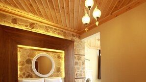 Un dormitorio con techo de madera, pared de piedra, espejo redondo, cama con ropa blanca y una mesita de noche.