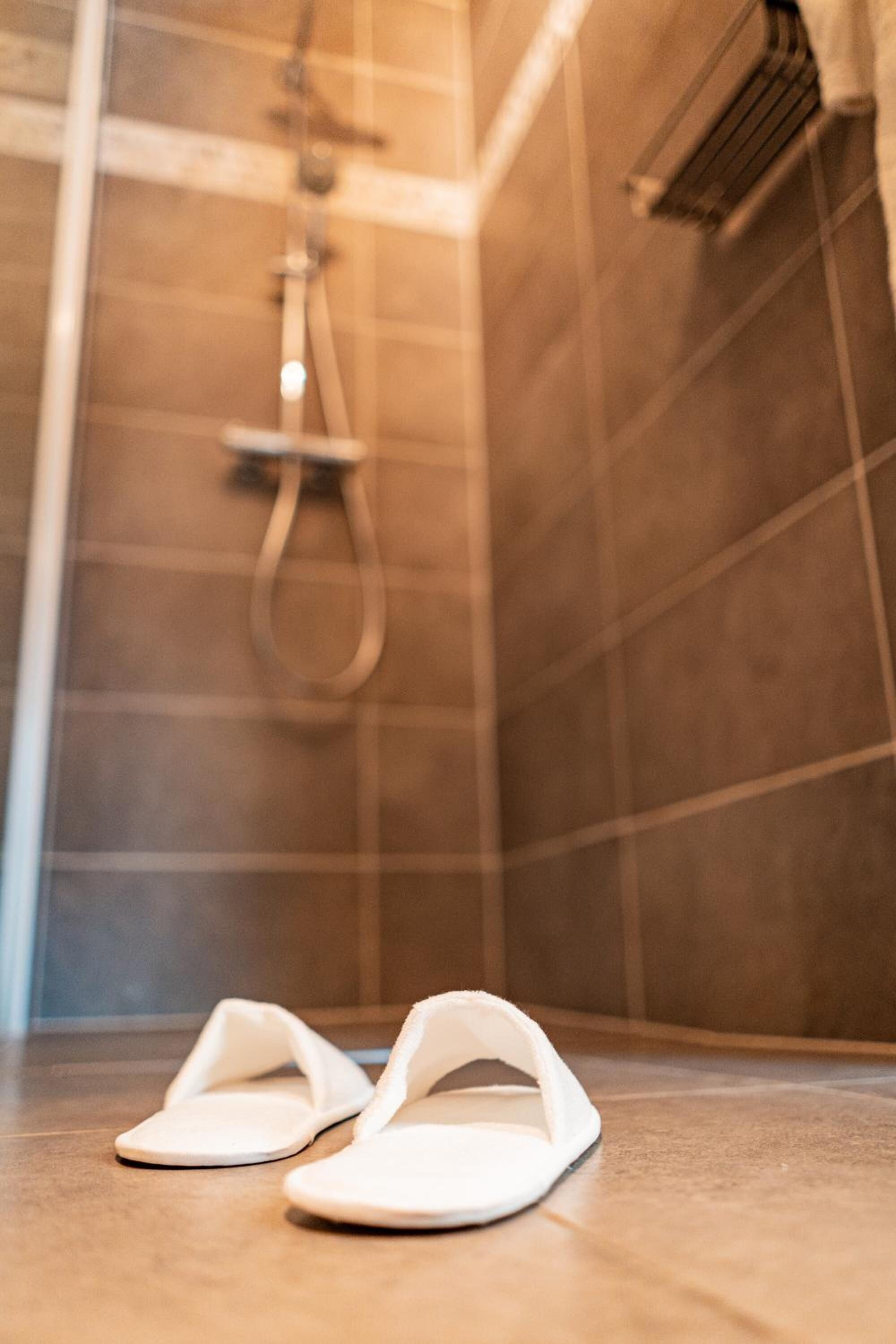 Een paar witte pantoffels op een betegelde vloer in een badkamer met een douche en een handdoekrek.
