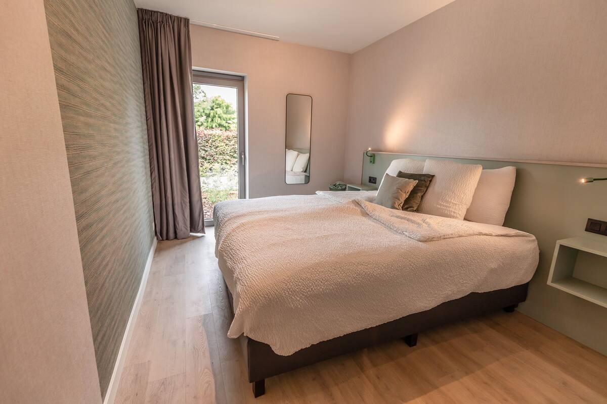A modern bedroom with a large bed, a bedside table, a mirror, and a view of the outdoors.