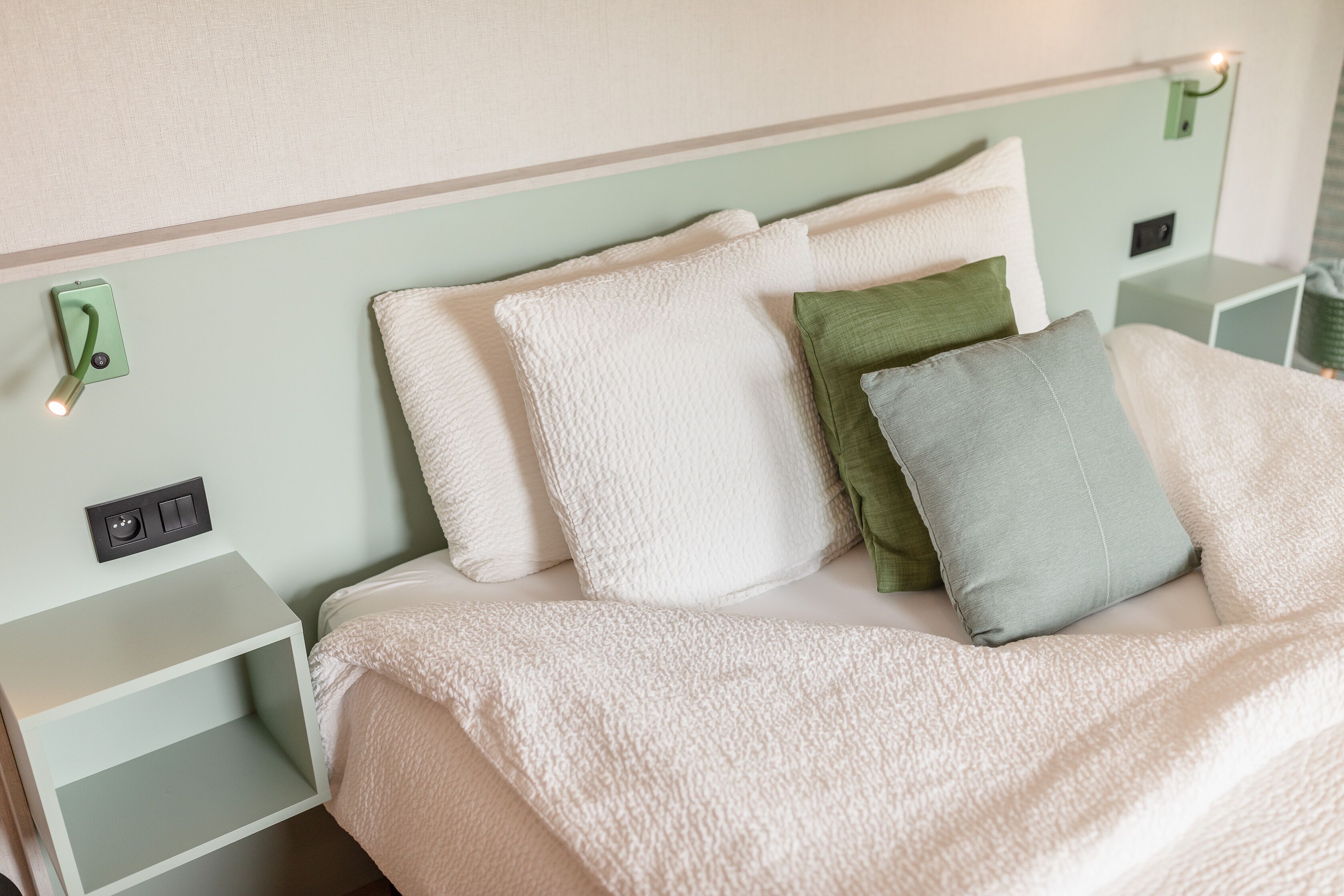 Una cama bien hecha con almohadas blancas y verdes, una manta blanca y una mesita de noche verde con una lámpara.