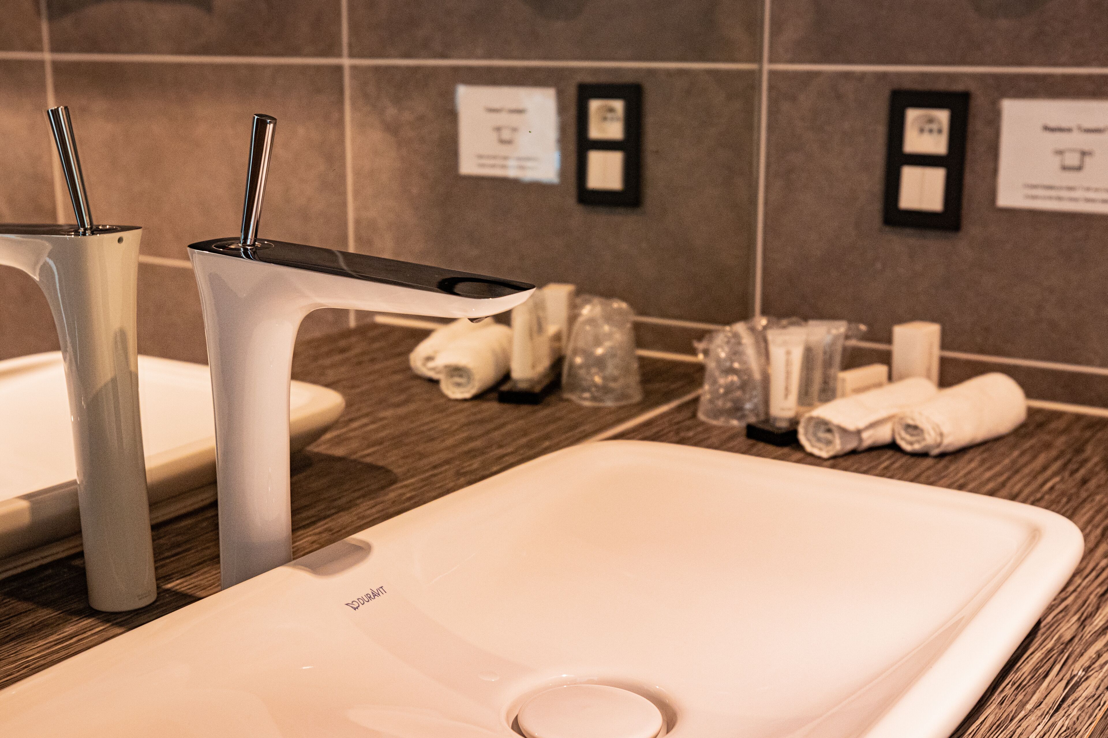 A modern bathroom with a large sink, two faucets, and a mirror. There are toiletries and towels neatly arranged on the counter.