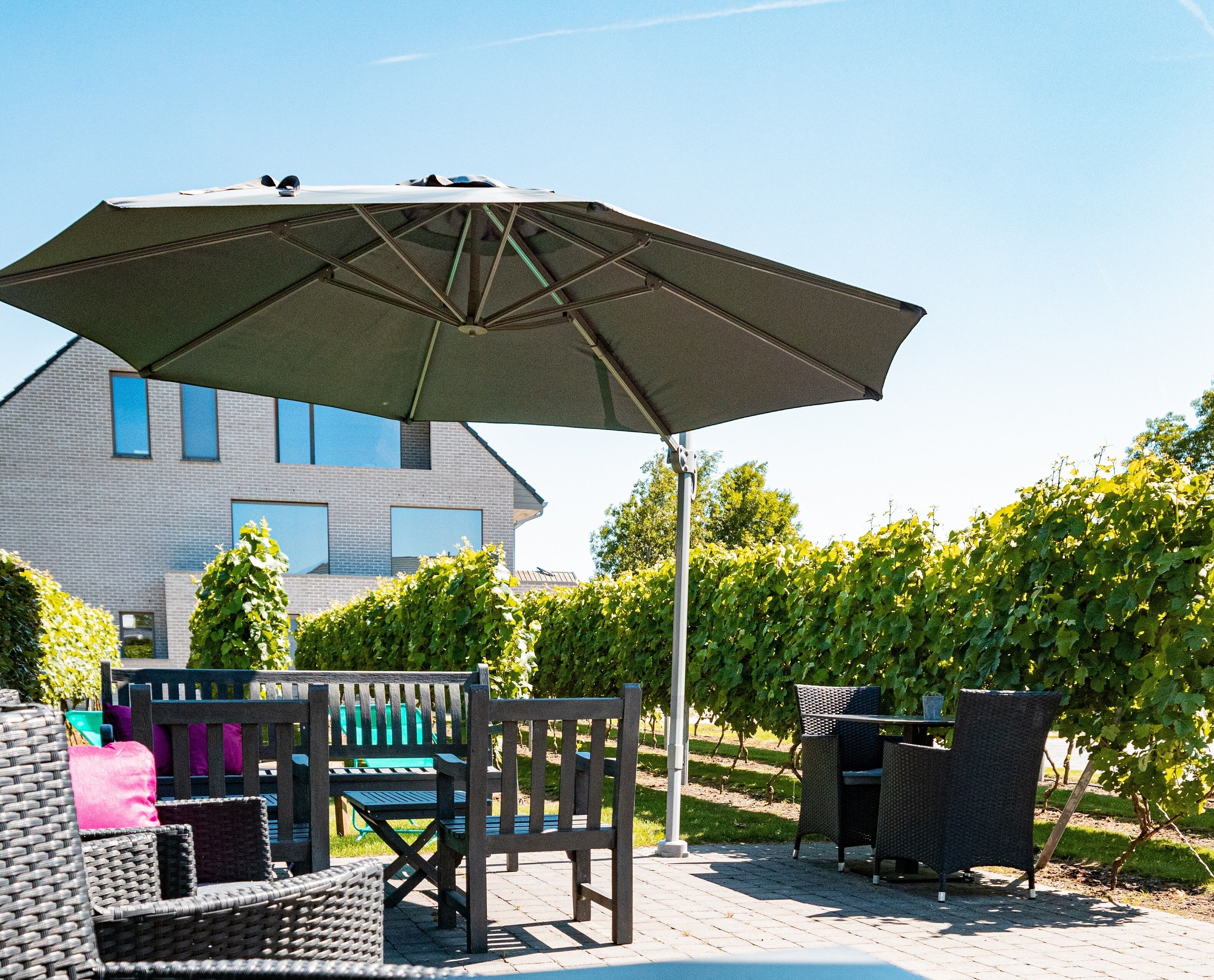 Une terrasse extérieure aménagée avec des meubles en osier, un grand parasol et une vue sur une maison et un vignoble en arrière-plan.