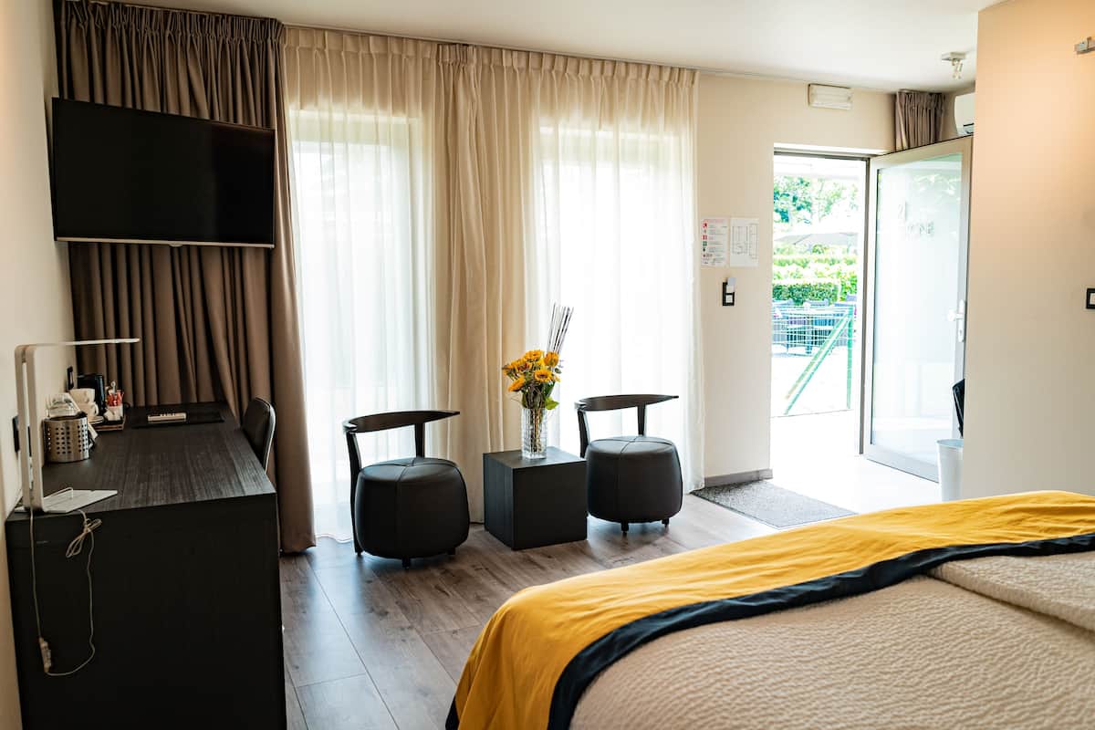 A hotel room with a bed, a desk with a computer, a TV, two ottomans, a vase with flowers, and a view of a garden through the door.