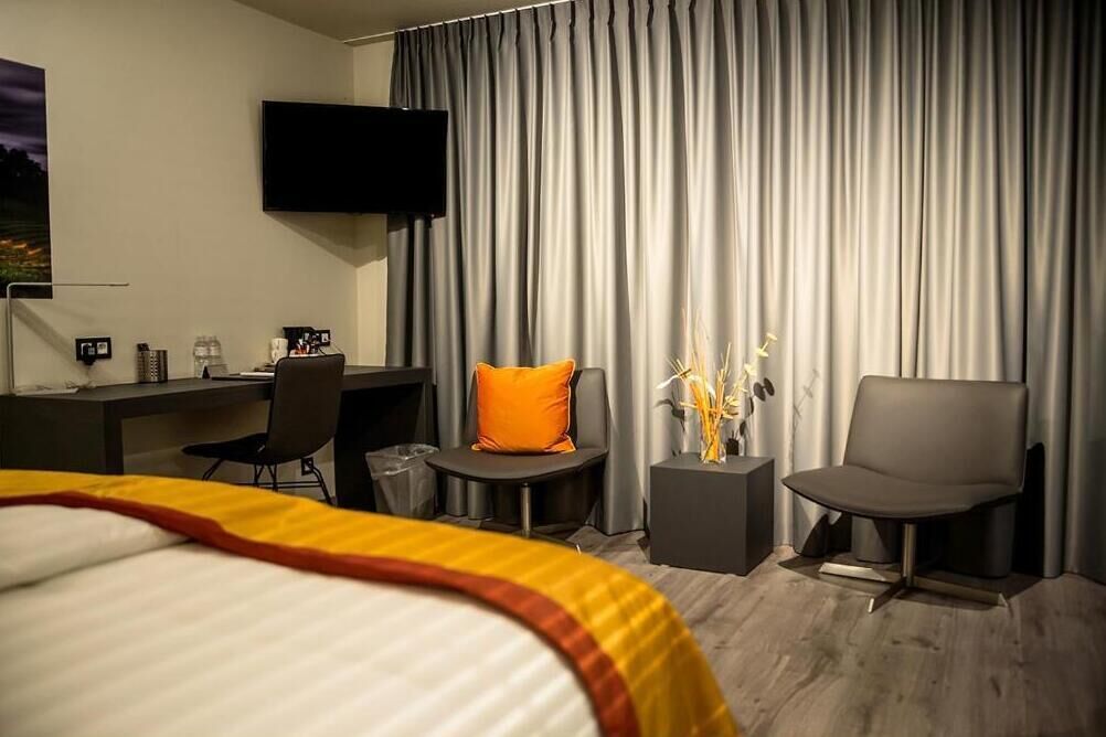 A hotel room with a bed, a desk with a chair, a TV mounted on the wall, and a large window with curtains.