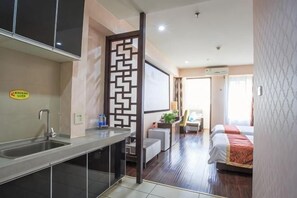 A hotel room with a kitchenette, a bed with a red blanket, a desk with a chair, and a view of the outdoors.