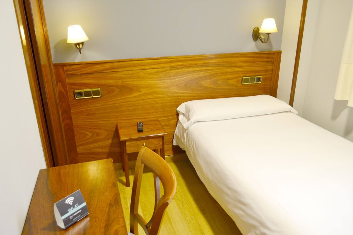 A hotel room with a wooden headboard, a single bed, a wooden nightstand with a remote control, and a chair.