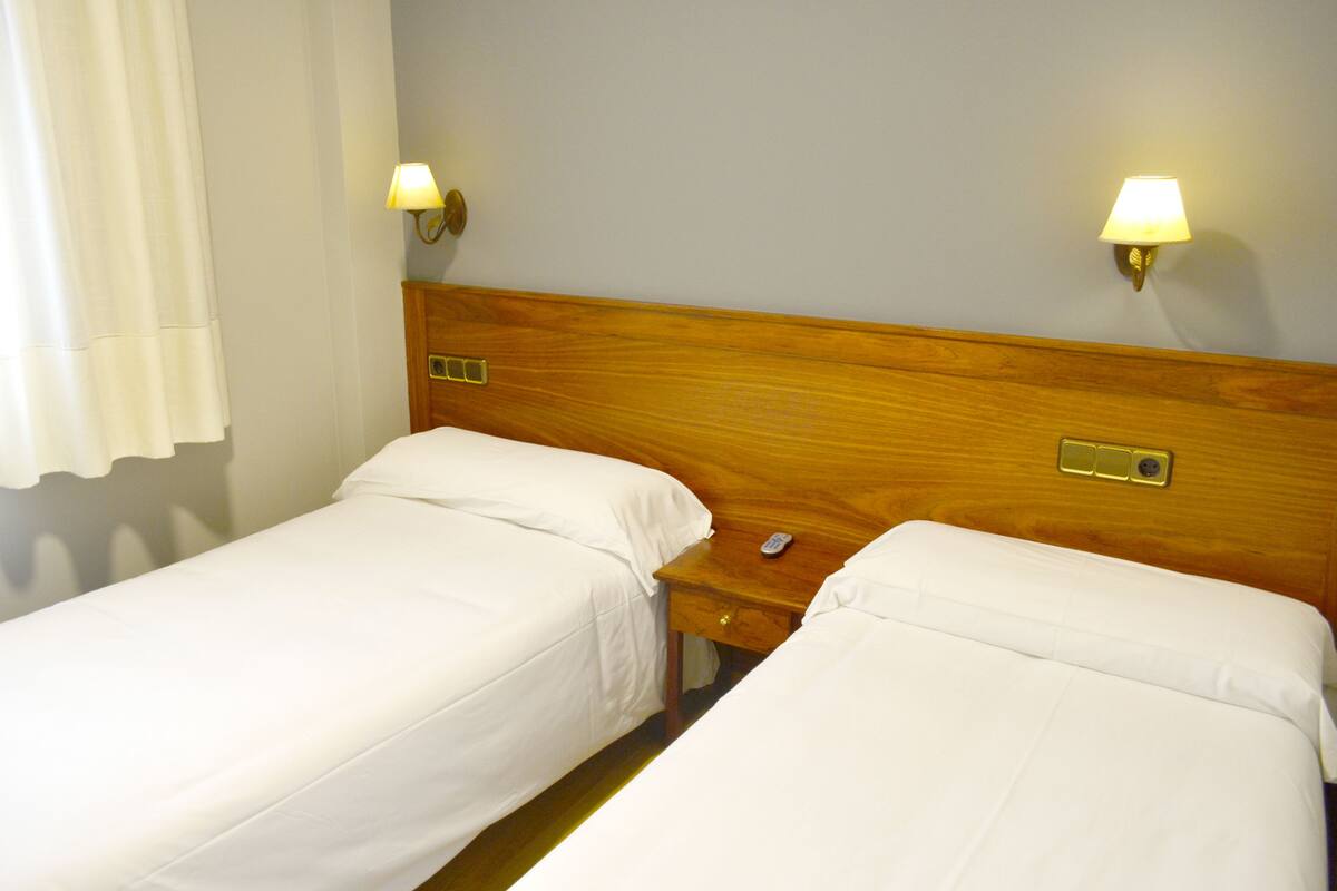 A hotel room with two single beds, wooden headboards, bedside tables with lamps, and a nightstand with a remote control.