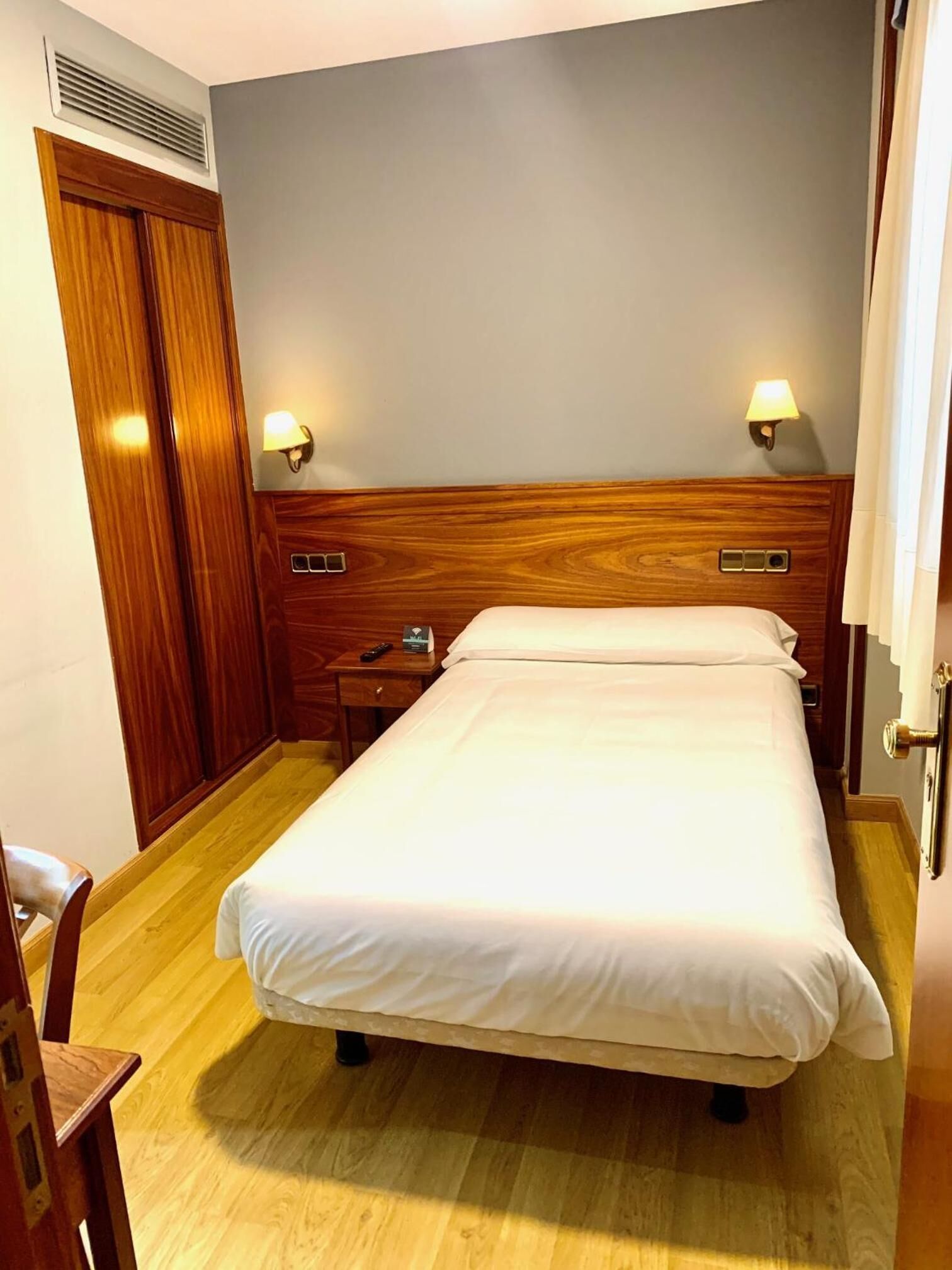 Habitación de hotel con cabecera de madera, cama con sábanas blancas, mesita de noche con lámpara y armario de madera.