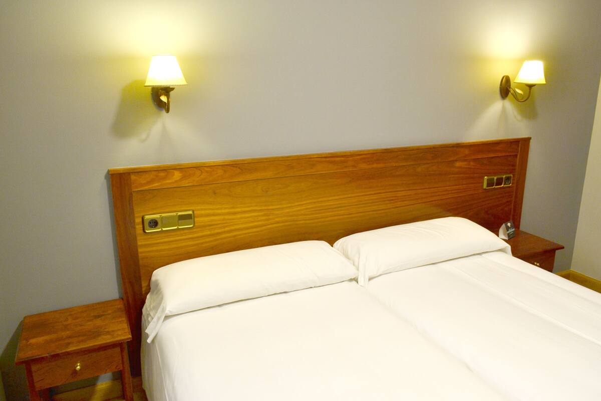 A double bed with white linens, wooden headboard, two bedside tables with lamps, and a nightstand with a drawer.