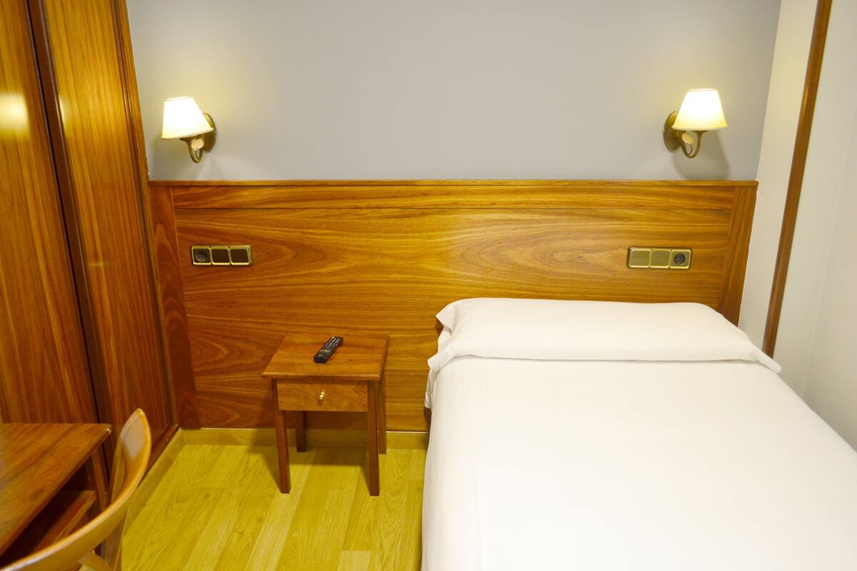 A hotel room with a wooden headboard, two bedside lamps, a small wooden nightstand with a remote control, and a bed with white bedding.