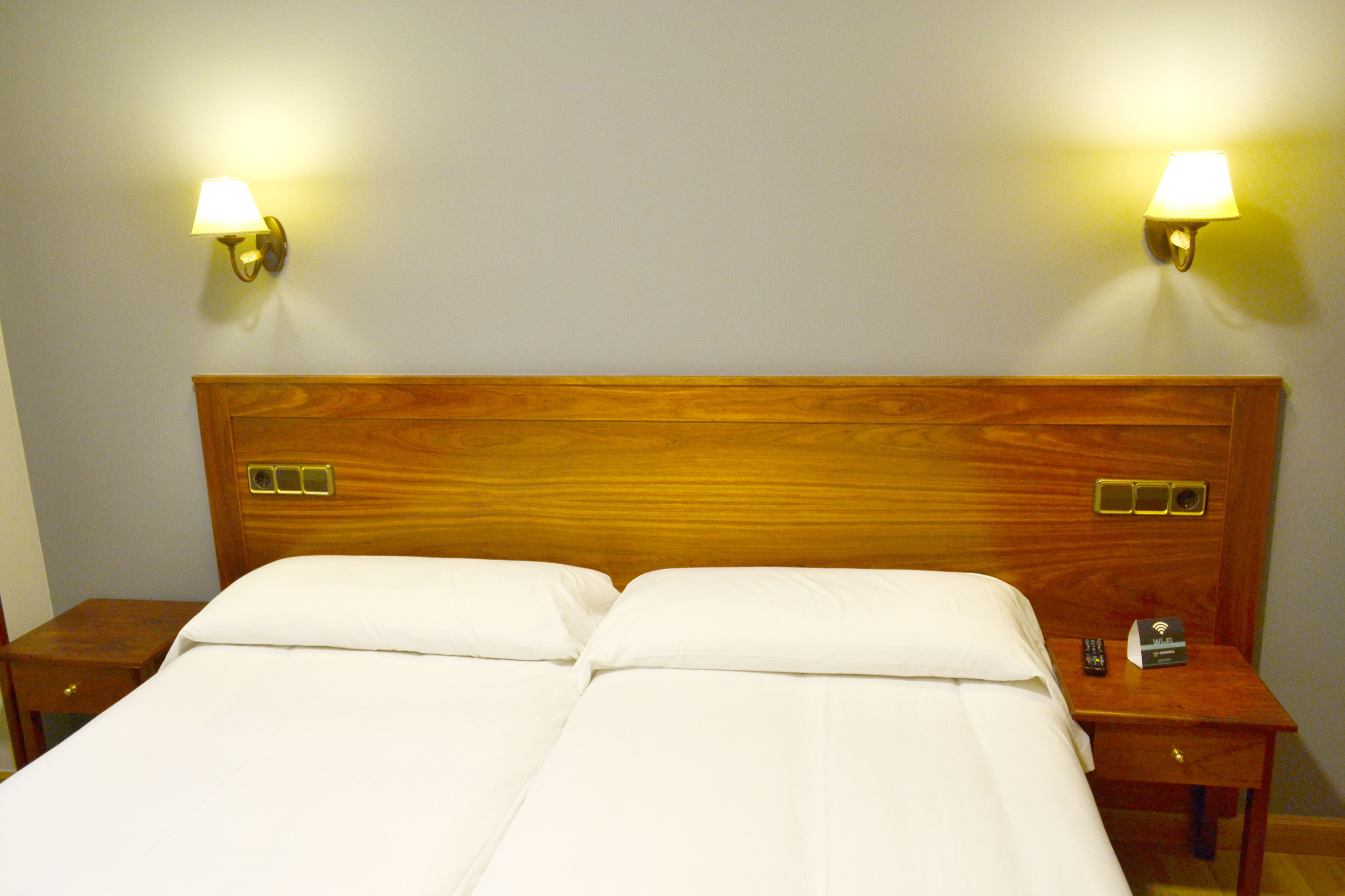 Habitación de hotel con cabecera de madera, dos lámparas de noche, una mesita de noche con control remoto y un pequeño box, posiblemente un minibar o artículos de servicio.