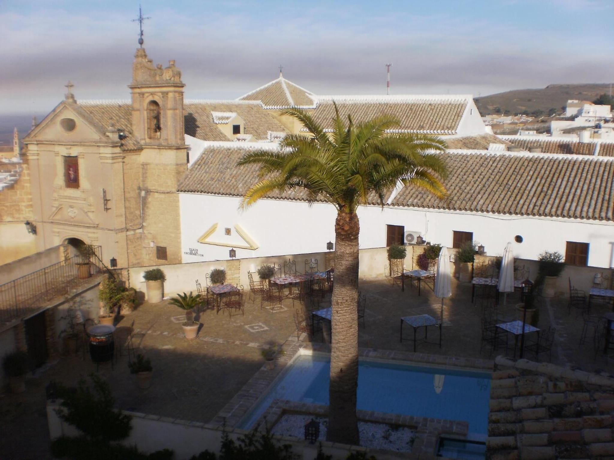 A courtyard with a pool, palm trees, and traditional architecture including a church and tiled roofs.