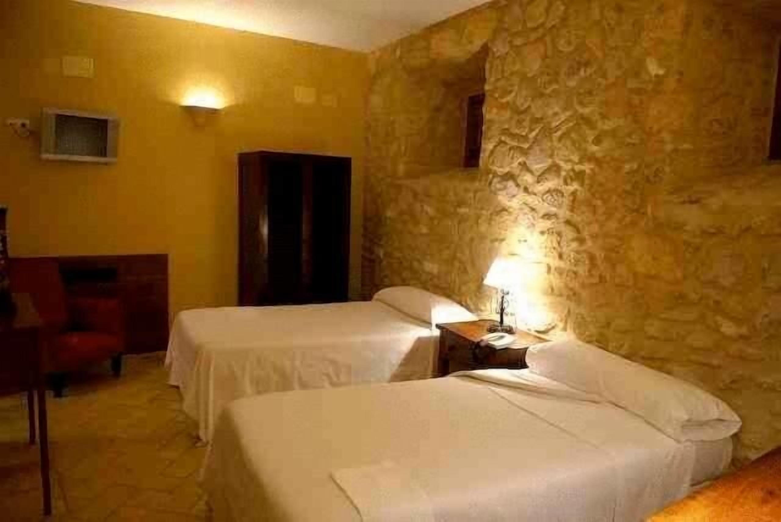 A room with two beds, a wooden wardrobe, a television, and a stone wall.