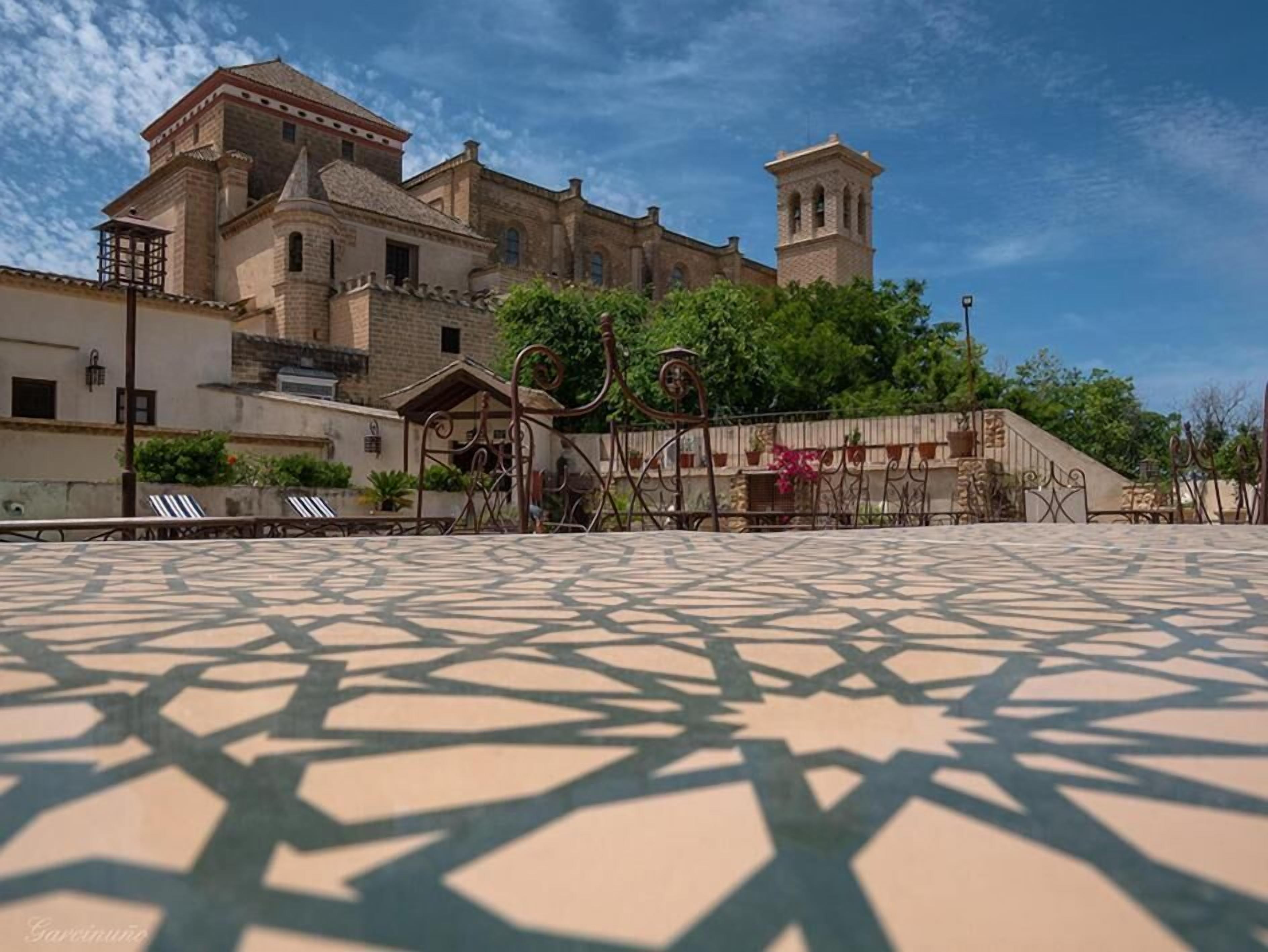 Une cour avec un sol décoré de motifs, un bâtiment doté d’un clocher et une pergola couverte de plantes grimpantes.