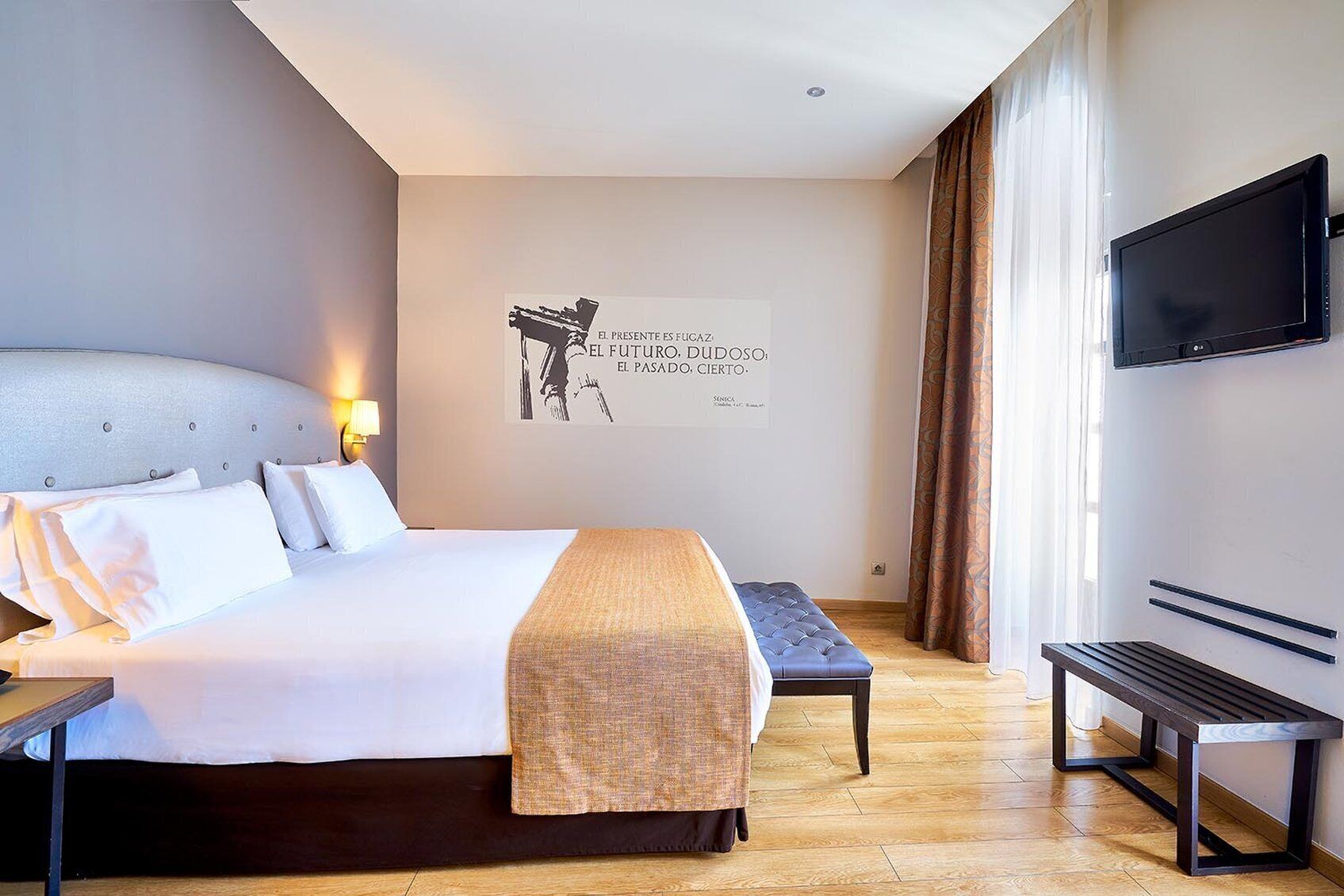 Una habitación de hotel moderna con una cama grande, un banco, un televisor de pantalla plana y una frase motivadora en la pared.