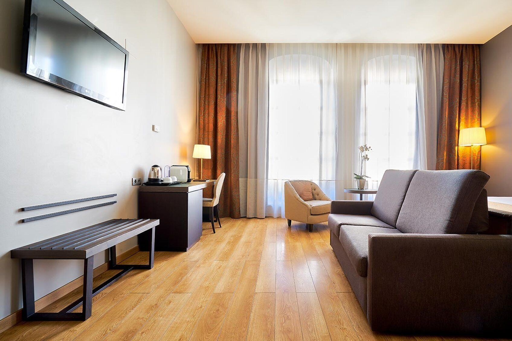 A hotel room with a flat-screen TV mounted on the wall, a desk with a chair, a sofa, a bench, and a small table with a lamp and an orchid.
