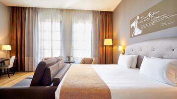 Habitación de hotel con una cama grande, un sofá, un escritorio y una lámpara. Se ve una ventana con cortinas y un cuadro en la pared.