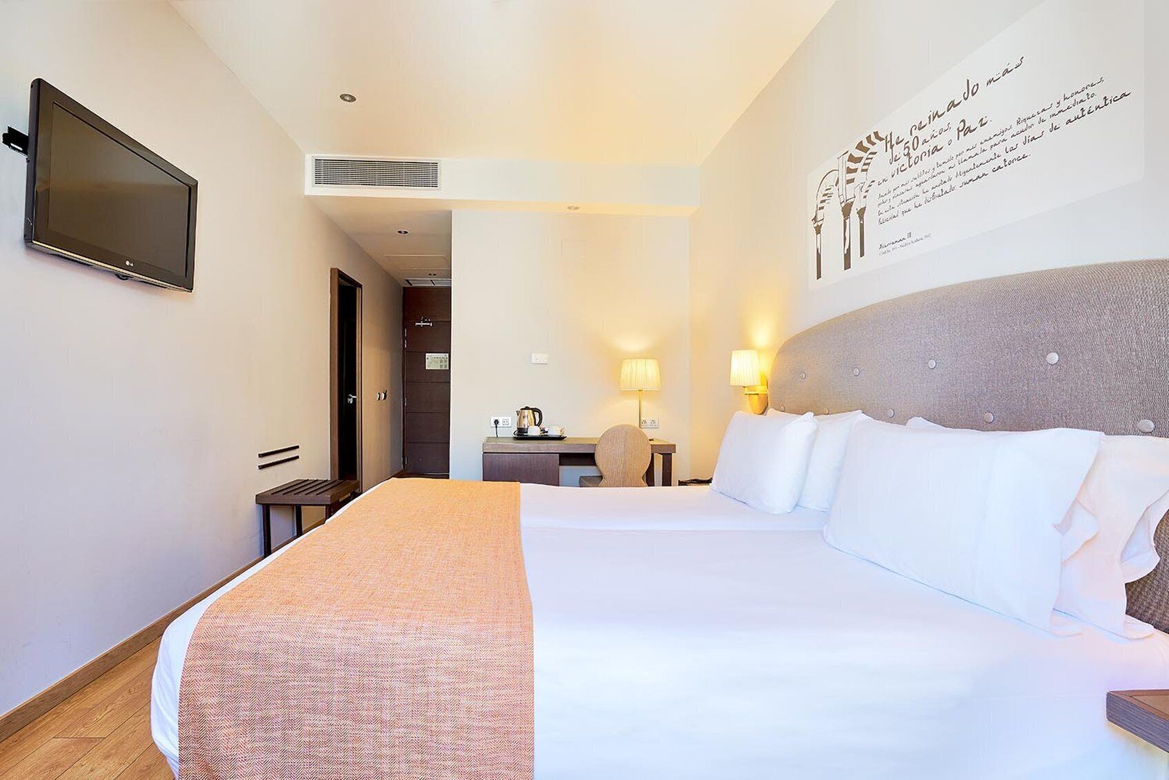 A hotel room with a large bed, a flat-screen TV mounted on the wall, a desk with a lamp, and a wall-mounted artwork.