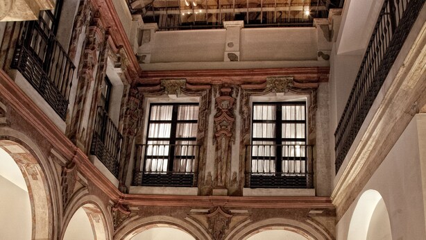 Un vestíbulo espacioso con techo alto, columnas ornamentales y piso a cuadros. Hay varias zonas de estar con mesas y sillas.