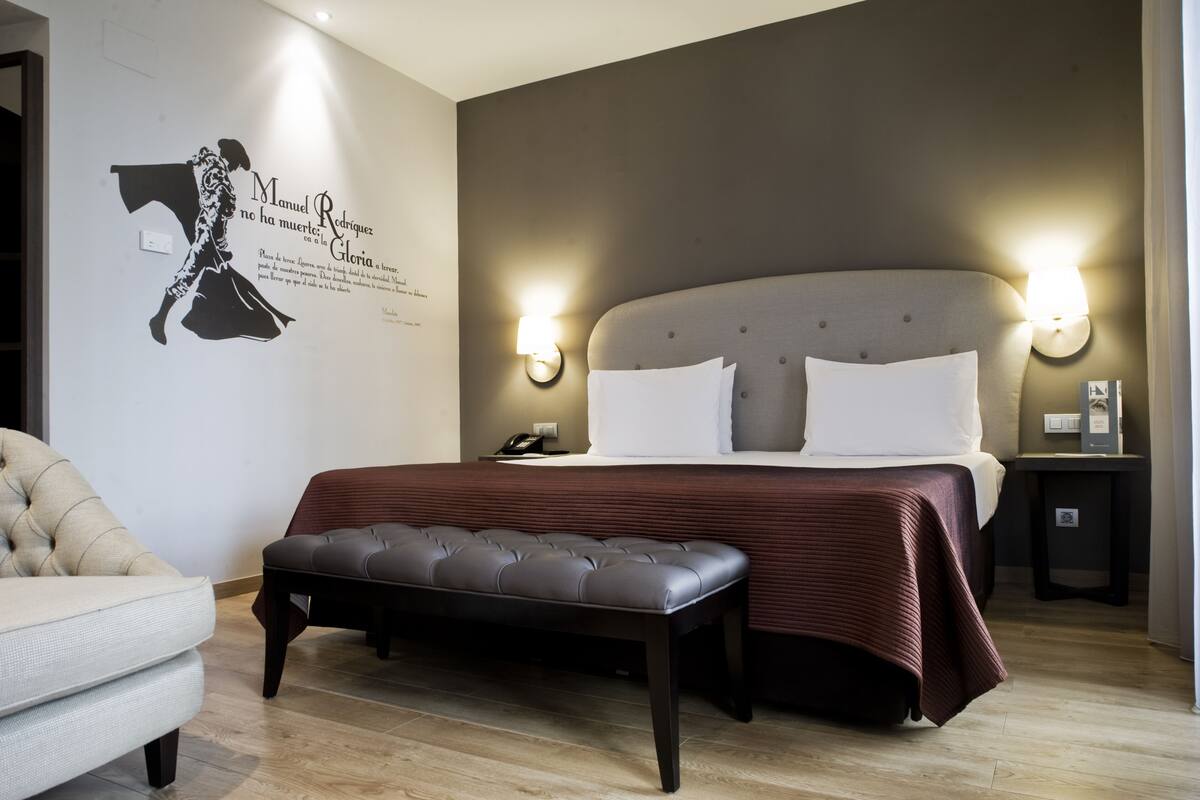 Un dormitorio con una cama grande, dos lámparas de noche, un banco, una silla y un adorno en la pared.
