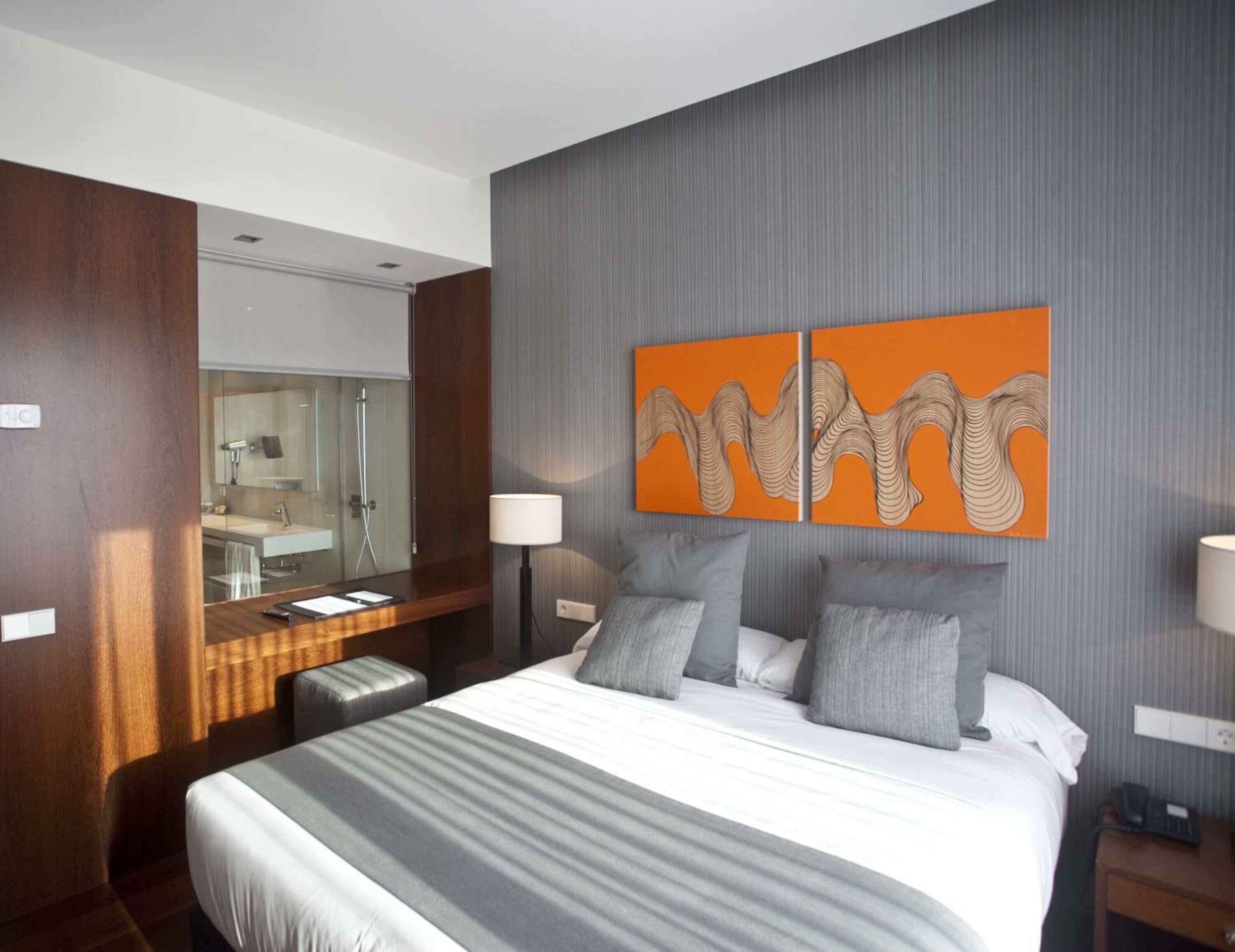 Une chambre d’hôtel moderne avec un grand lit, un bureau en bois, un miroir et deux œuvres d’art aux couleurs orangées accrochées au mur.