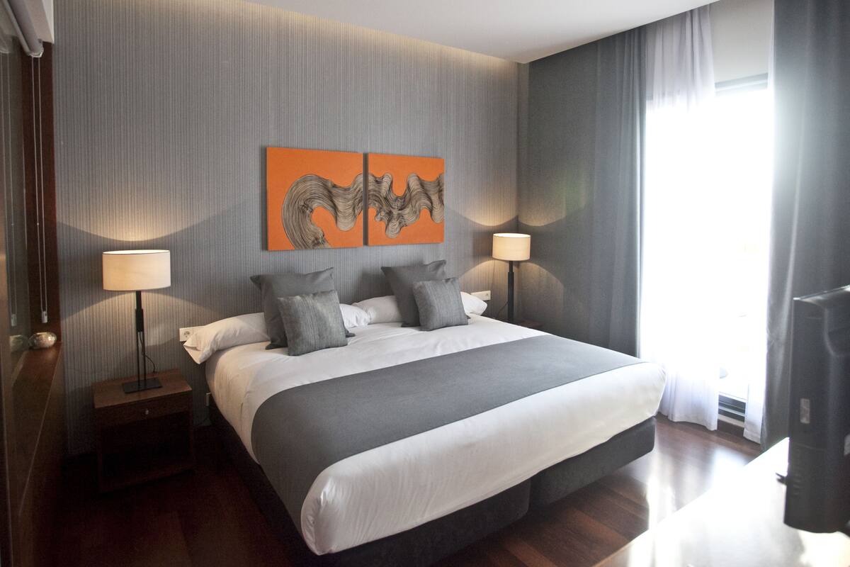 A modern hotel room with a large bed, two bedside lamps, a flat-screen TV, and a window with curtains.