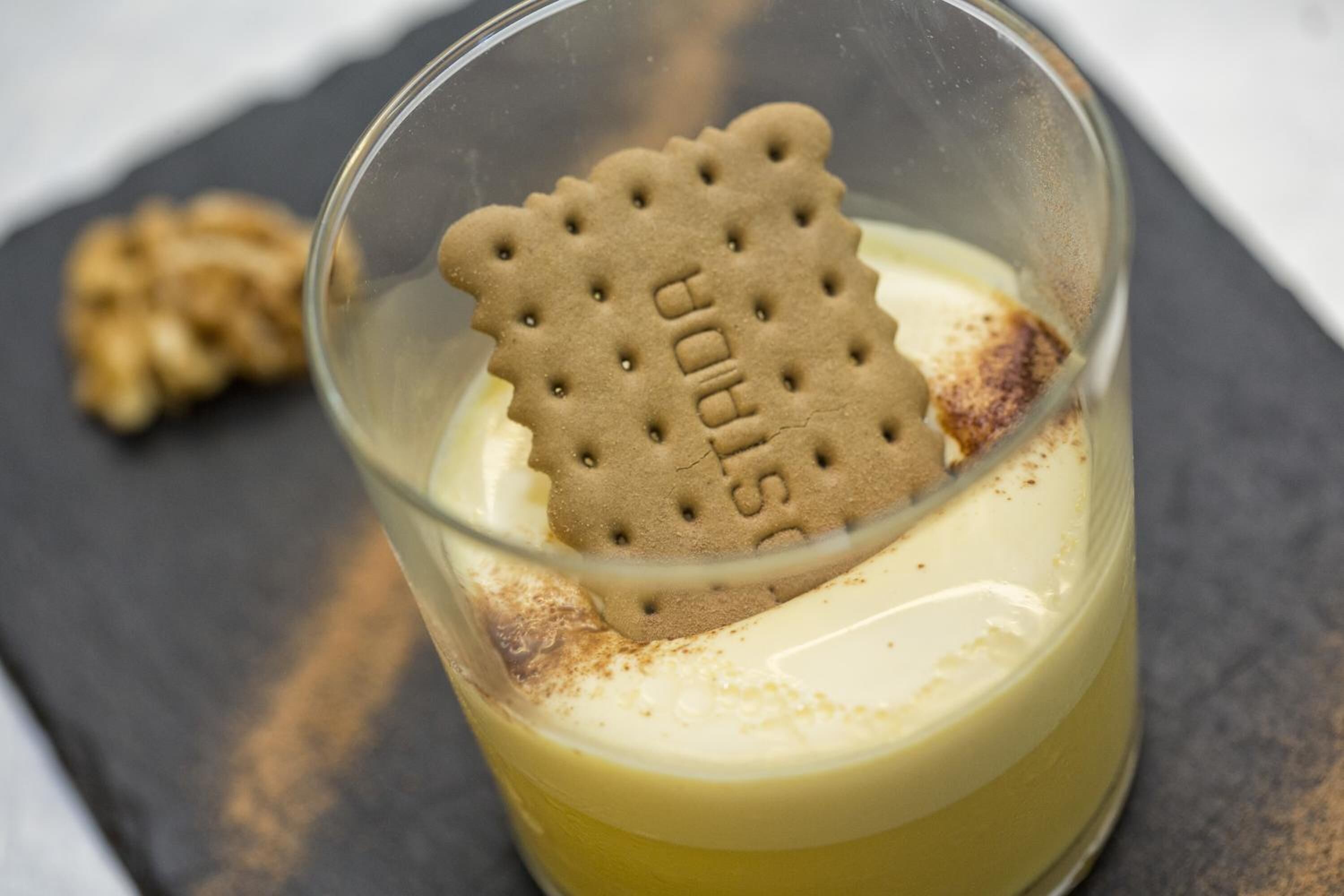 A dessert in a glass with a cookie on top, a nut in the background, and a dark surface beneath.