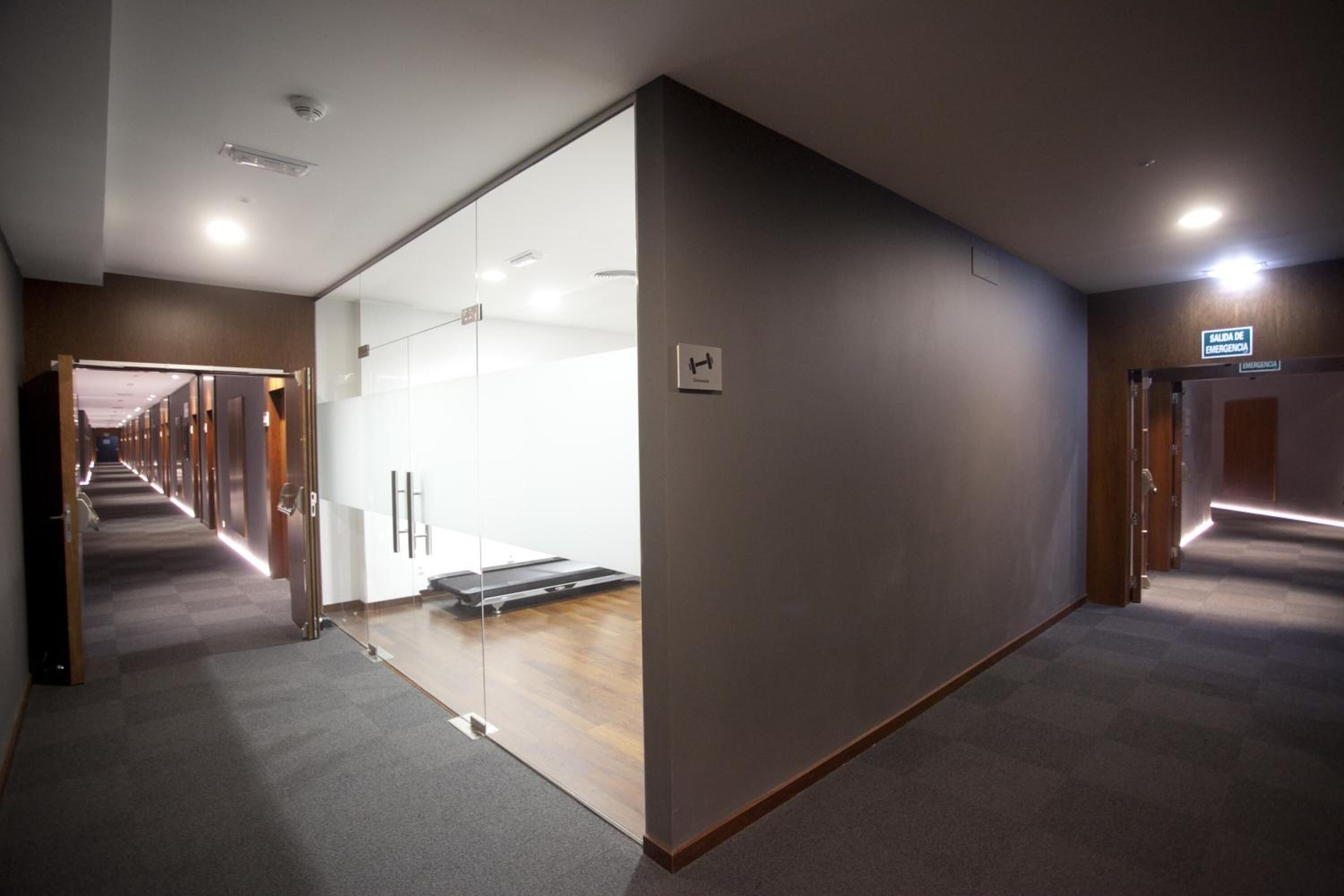 Et hotelkorsgang med glasvæg, løbebånd og skilt der viser vejen til fitnesscenteret.