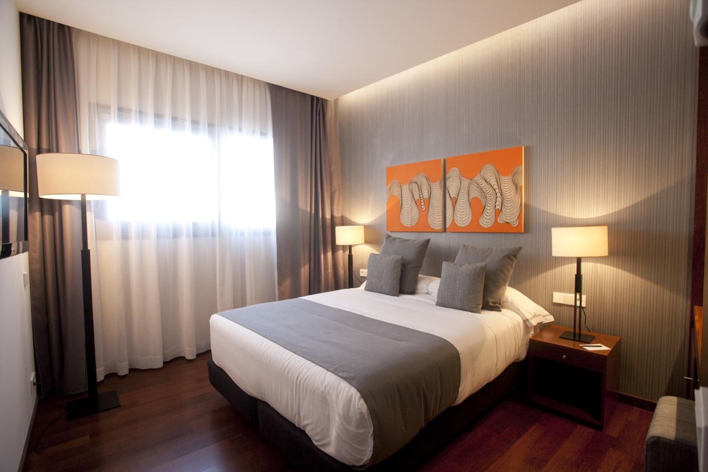Quarto de hotel com uma cama grande, duas luminárias de cabeceira, uma mesa de cabeceira e uma janela com cortinas.
