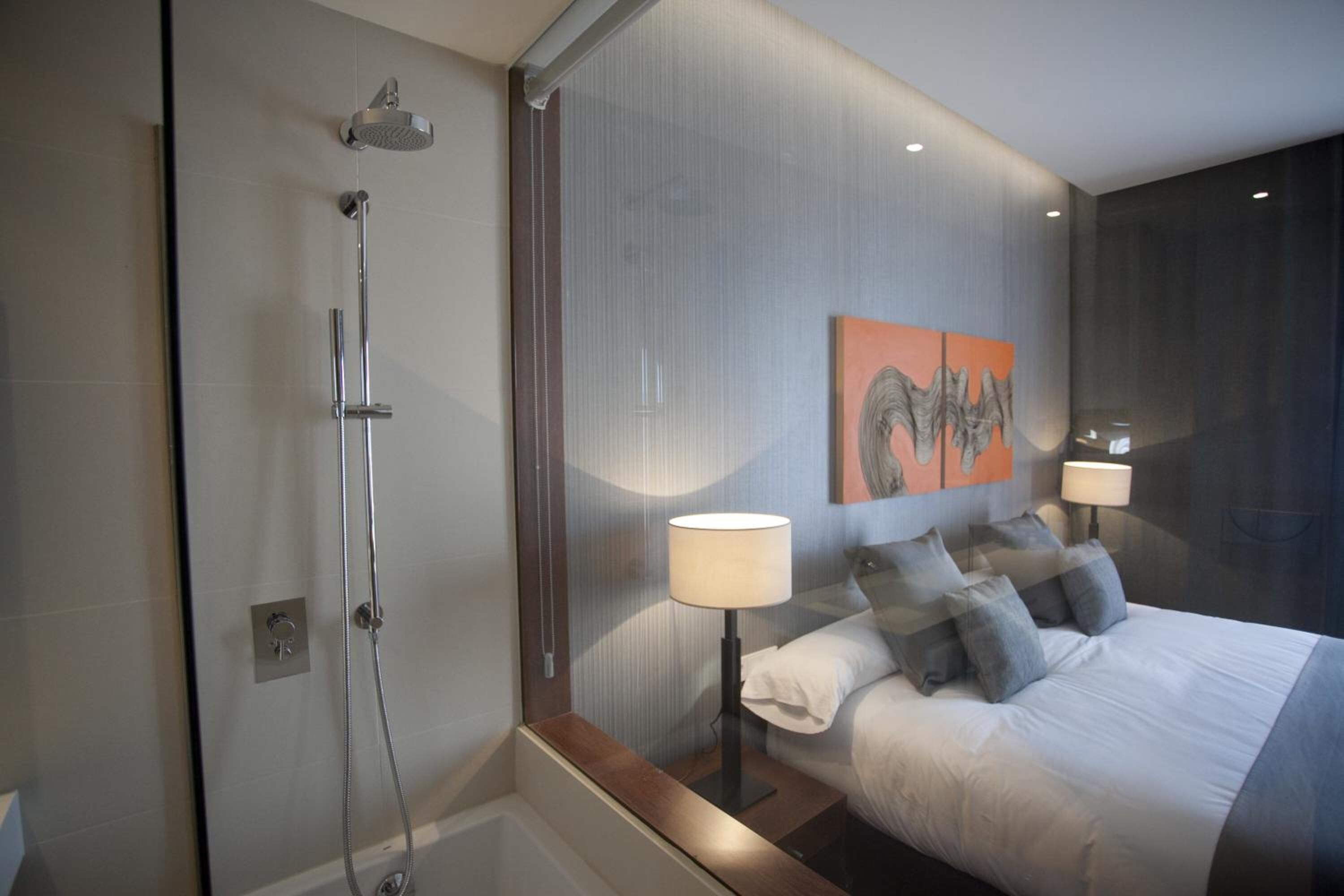 Una habitación de hotel moderna con cabina de ducha de vidrio, una cama con muchas almohadas y dos lámparas de noche.