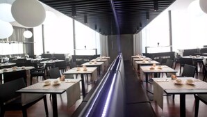 Un restaurante moderno con mesas y sillas largas, ventanas amplias y un elemento de iluminación central.