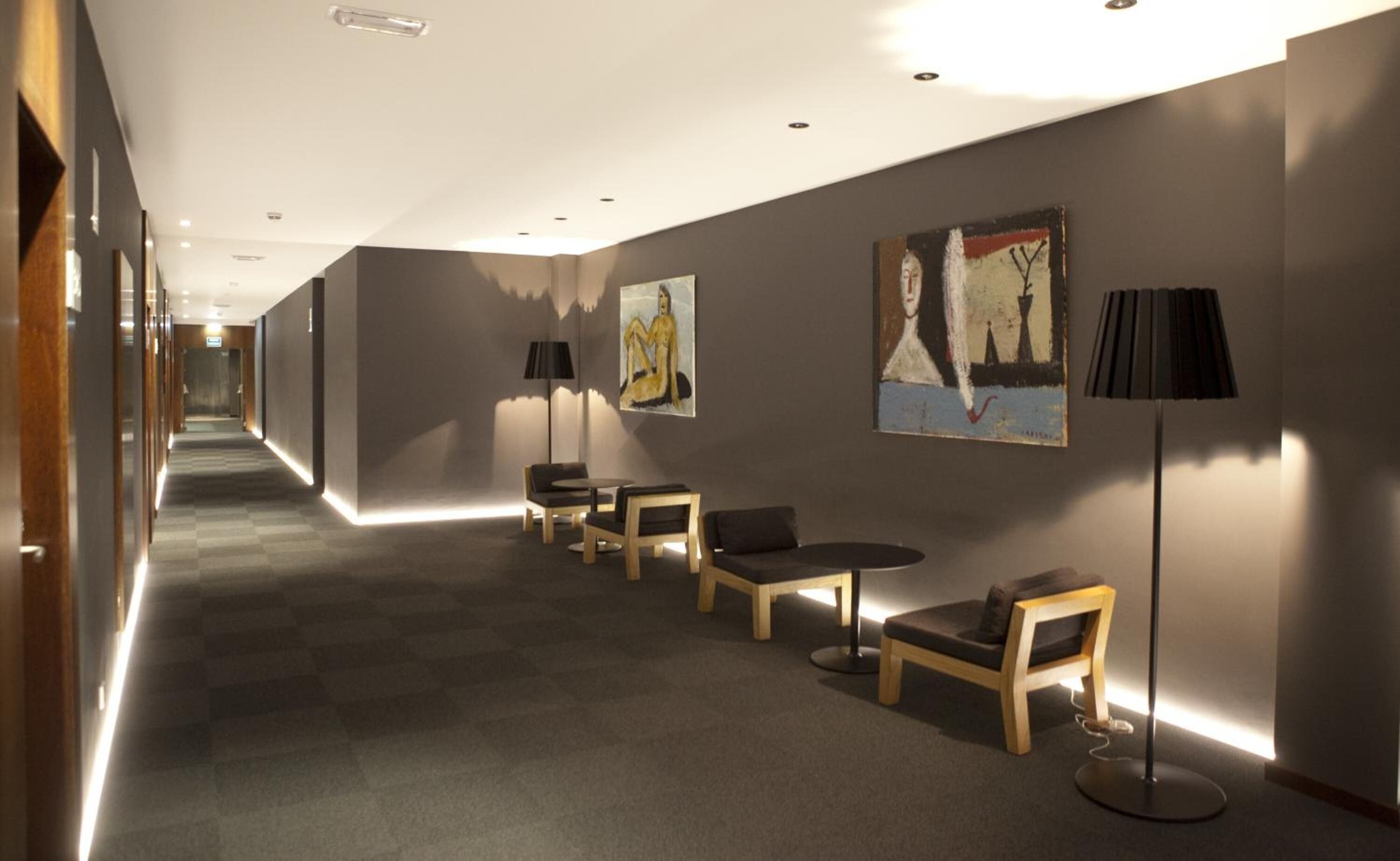 Un couloir doté d’un sol recouvert de moquette, de chaises en bois, d’une table et de deux tableaux accrochés au mur.