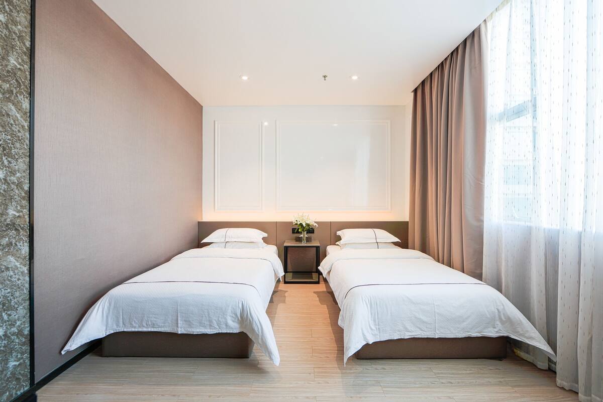 Une chambre d’hôtel avec deux lits, une petite table sur laquelle se trouve un vase, et une grande fenêtre avec des rideaux.