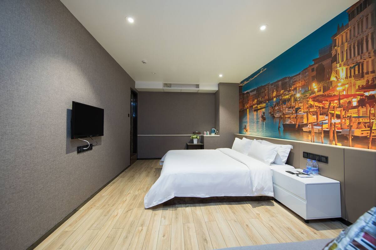 Une chambre d’hôtel moderne avec un grand lit, un téléviseur à écran plat fixé au mur, une petite table de chevet avec une bouteille, et une fresque représentant un paysage urbain sur le mur.