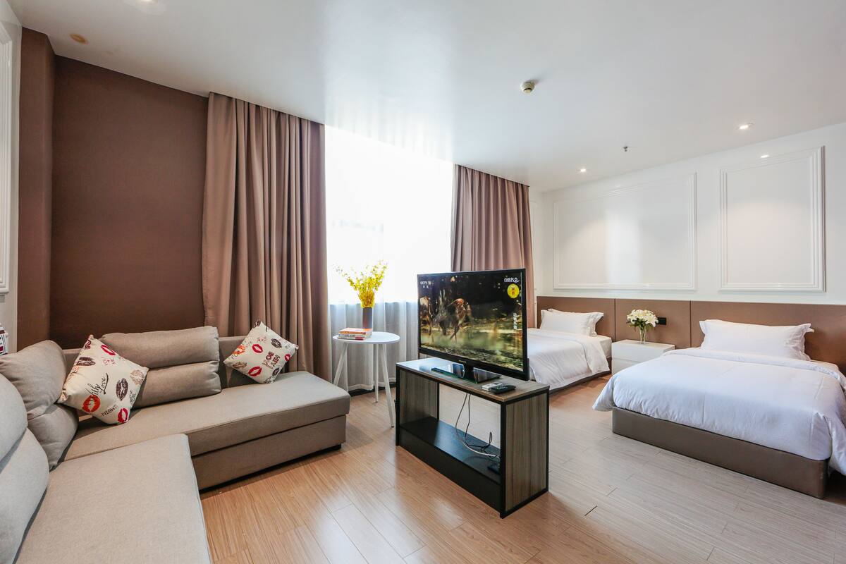 Une chambre d’hôtel moderne avec un canapé, un lit, un téléviseur posé sur un meuble et une petite table sur laquelle se trouve un vase.