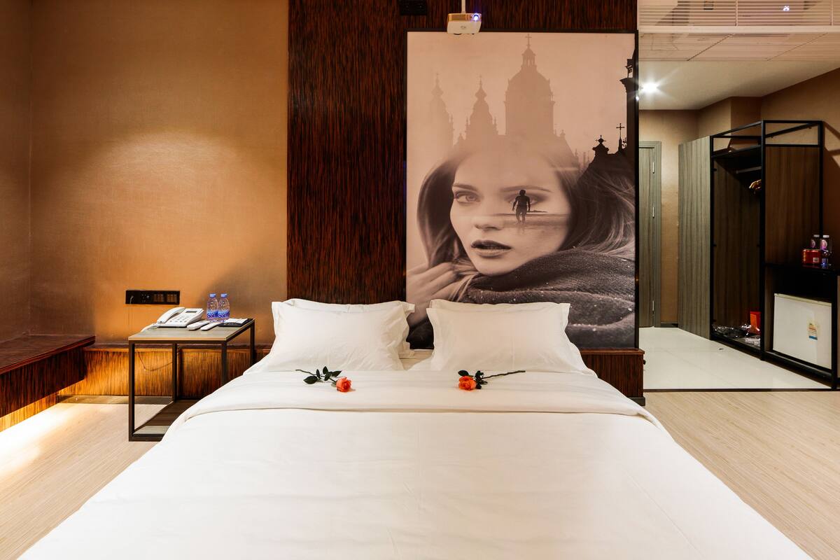 Une chambre d’hôtel avec un grand lit, une table de chevet avec un téléphone et des bouteilles d’eau, et une affiche représentant un bâtiment accrochée au mur.