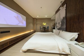 Una habitación de hotel moderna con una cama grande, un escritorio con dos sillas, una mesita con un florero y una pantalla grande en la pared.
