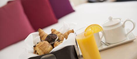 A breakfast tray with pastries, a glass of orange juice, and a teapot on a bed with pillows.