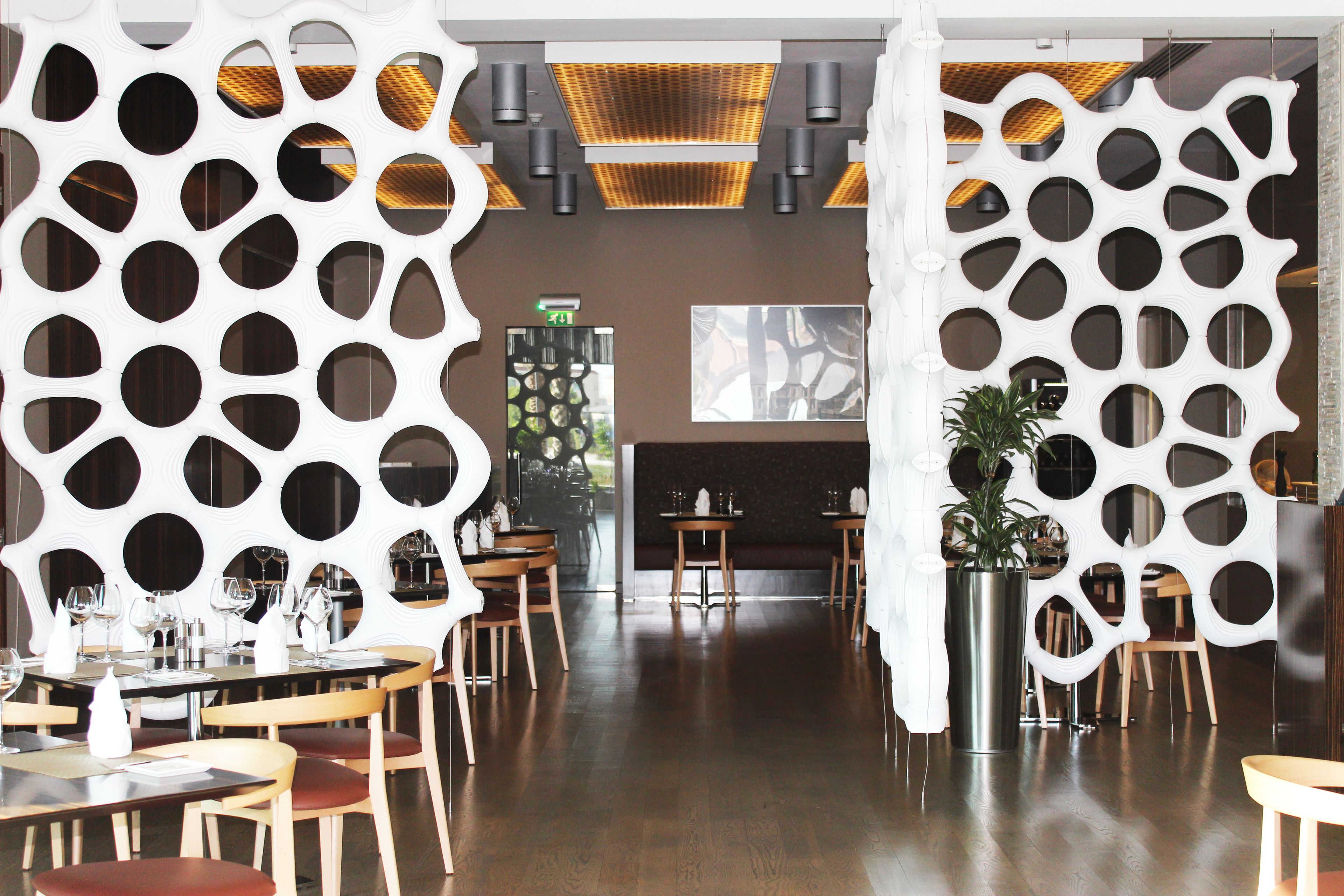 A modern restaurant interior with wooden tables and chairs, a central bar area, and decorative white panels with circular cutouts.