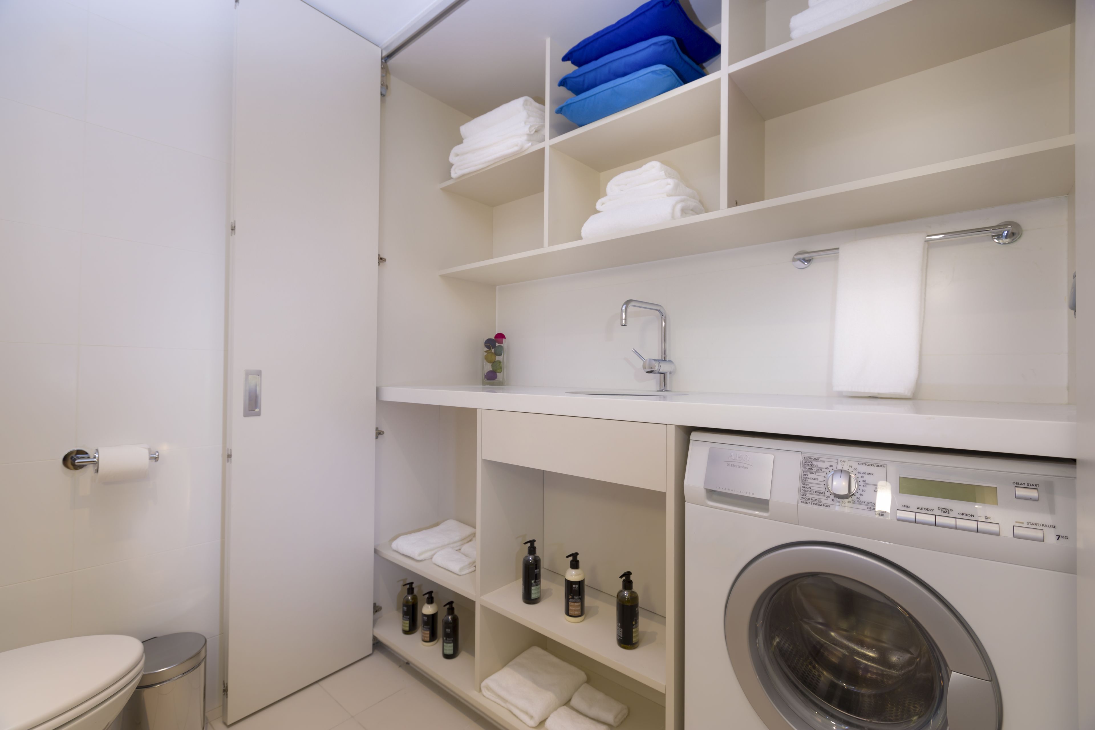 A compact bathroom with a toilet, a washing machine, and shelves with folded towels and toiletries.
