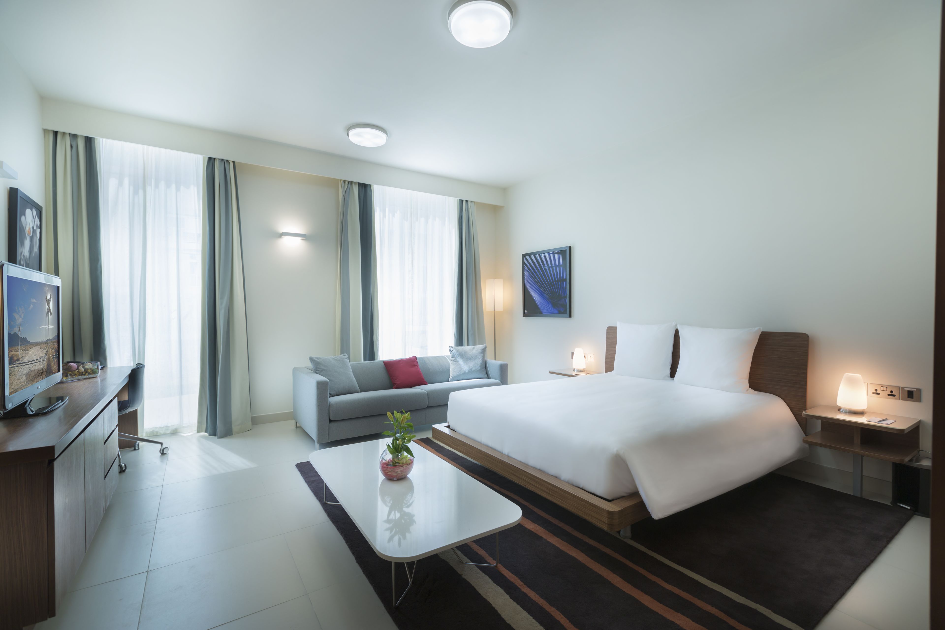 A modern hotel room with a large bed, a sofa, a desk with a computer, and a TV.