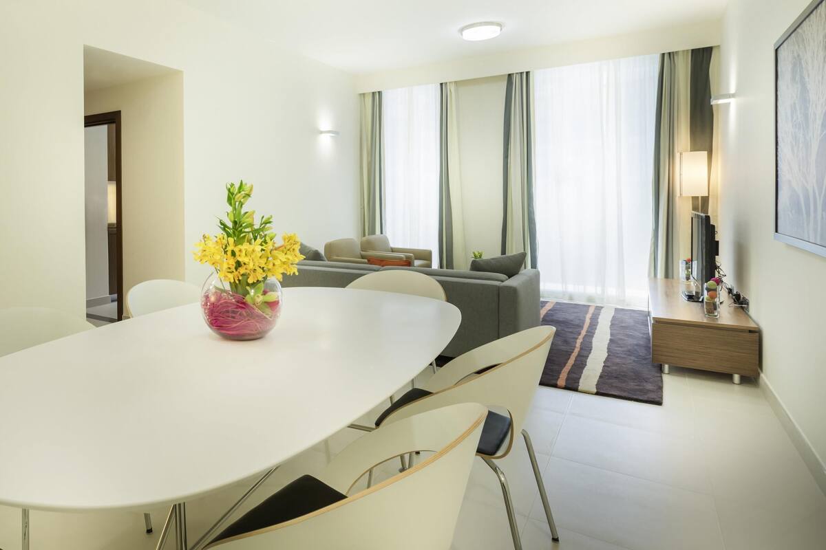 A modern living room with a white dining table, grey sofa, and a small side table with a lamp.