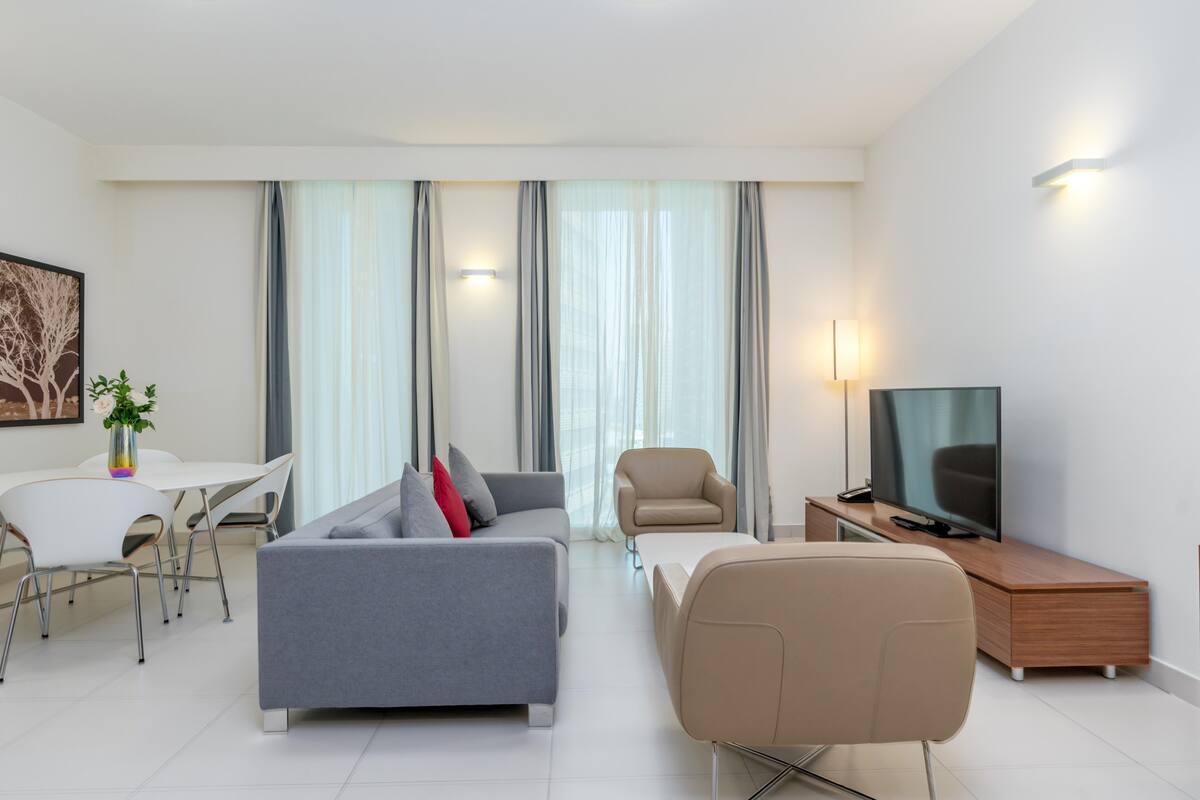 A modern living room with a grey sofa, beige armchair, and a TV on a wooden cabinet. There is a dining area with a white table and chairs, and a plant on the table.