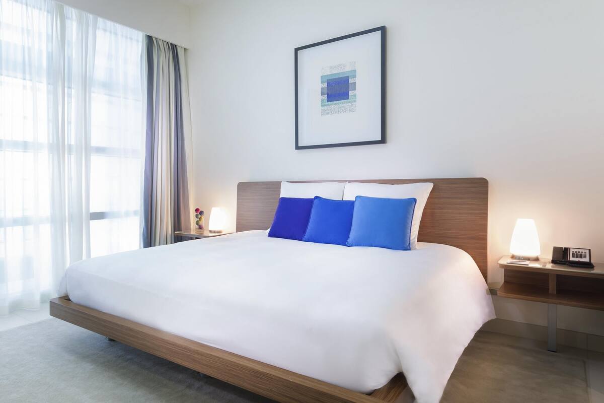 A hotel room with a large bed, a bedside table with a lamp, a framed picture on the wall, and a window with curtains.