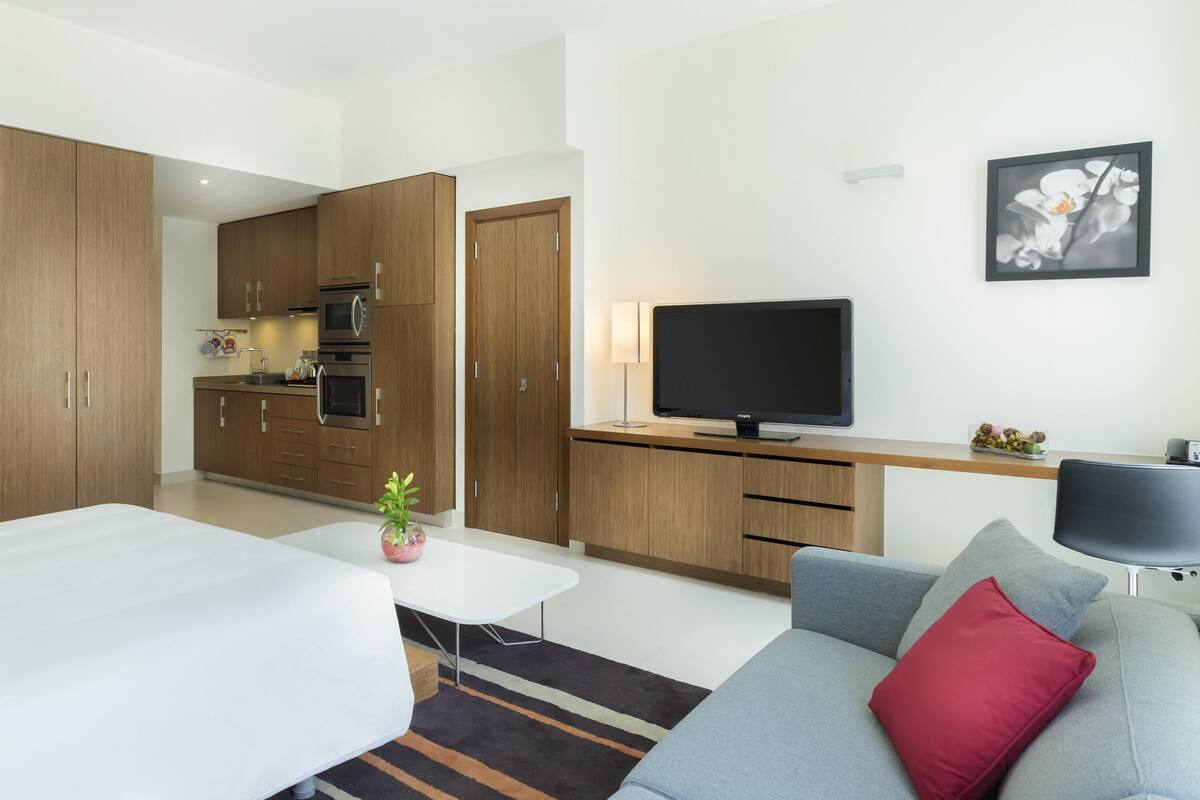 A modern hotel room with a kitchenette, a flat-screen TV, a sofa with a red pillow, and a striped rug.