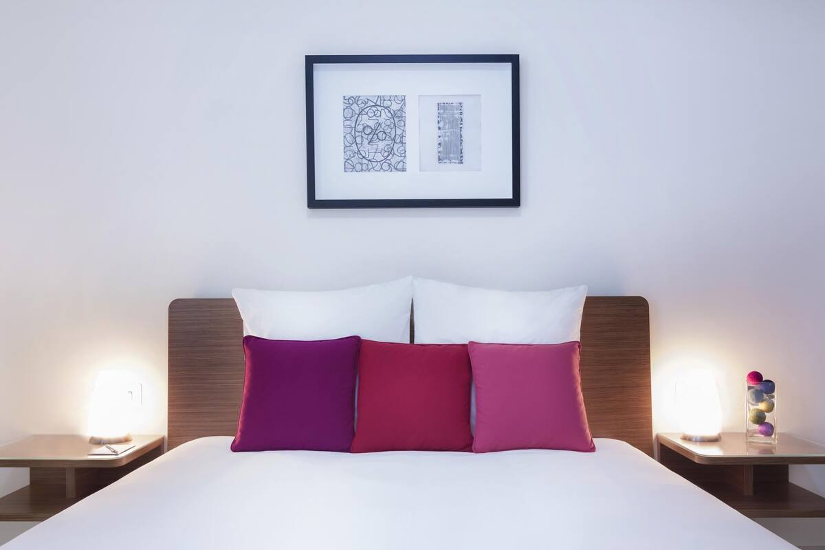 A neatly made bed with white and pink pillows, a framed artwork above it, and bedside tables with lamps and decorative items.