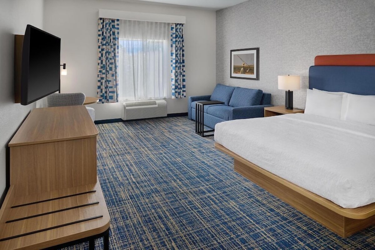 Habitación de hotel con cama, escritorio, televisión, sofá, lámpara y ventana con cortinas.