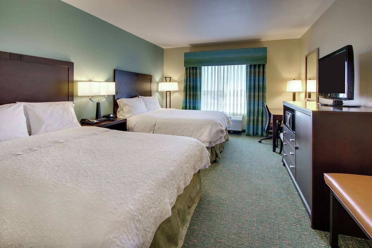 A hotel room with two beds, a desk, a television, and a window with curtains.