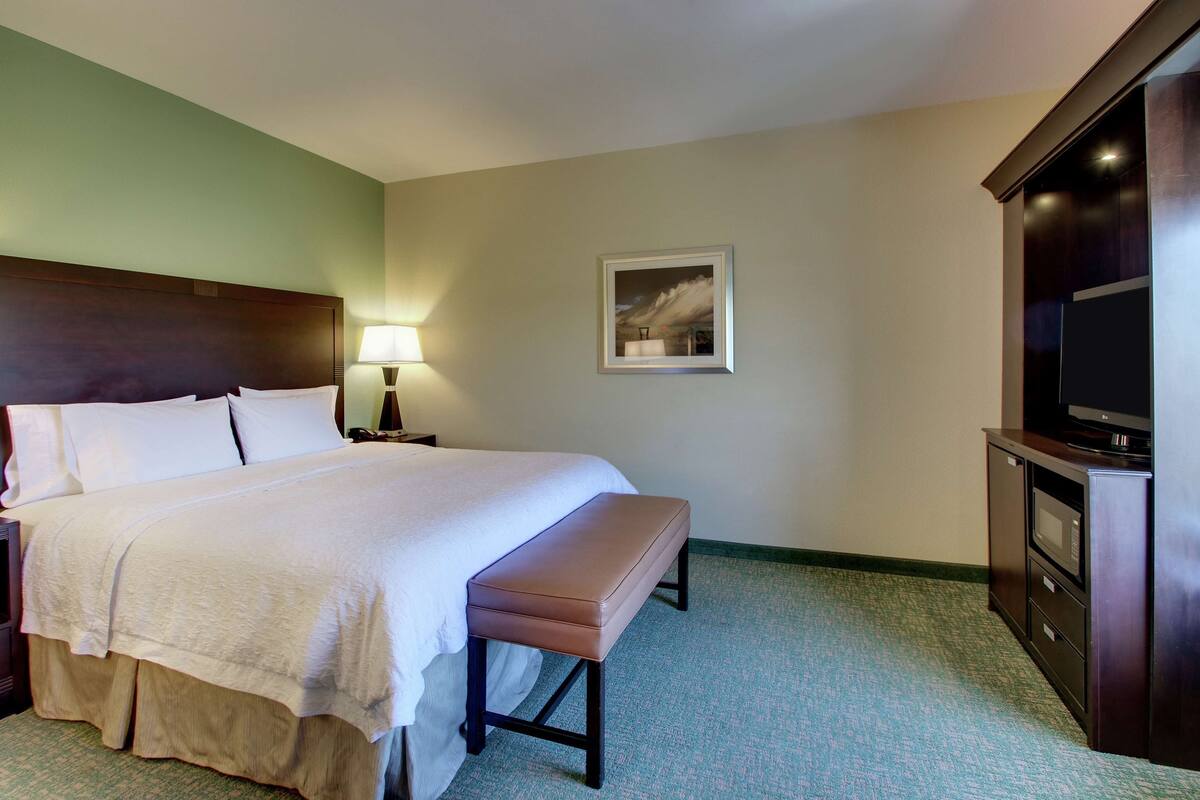 A hotel room with a bed, a bench, a TV, a lamp, and a framed picture on the wall.