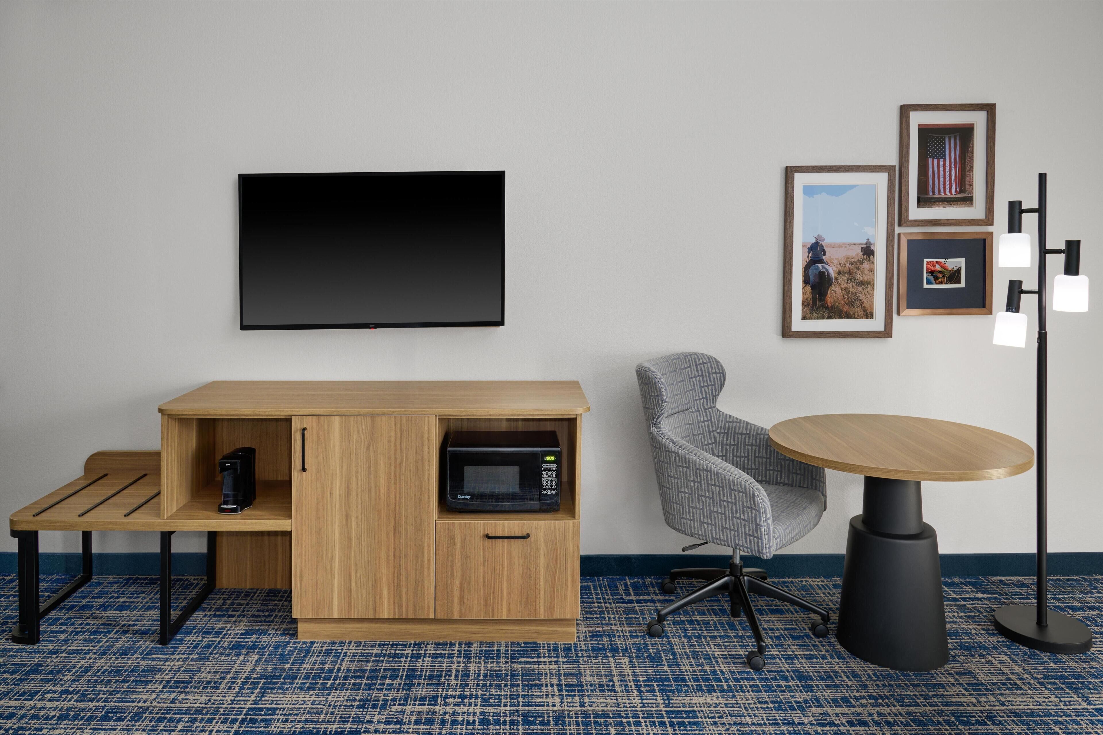A modern hotel room with a flat-screen TV, a wooden cabinet with a microwave, a gray office chair, a round table, and framed pictures on the wall.